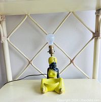 Mid century Blackamoor lady chalkware lamp shown unlit on chair with visible minor chip on feet area, light bulb socket attached above headwrap and yellow painted dress and headwrap.