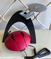Photo of the Star Trek themed telephone handset designed as the enterprise starship with black stand and red base, coiled cord visible.