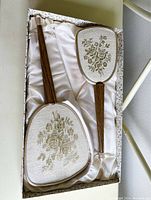 Vanity set in box showing the mirror and brush with embroidered fabric backs and wooden handles on cream satin lining.