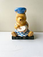 Front view of ceramic Winnie the Pooh cookie jar sitting on a book, wearing a blue chef hat and apron, holding a plate with honey dipper.