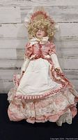 Full view of the Victorian-style doll dressed in pink and white lace dress with bonnet and gloves, showing detailed costume and face.