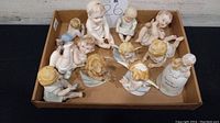 Top-down view of all eleven figurines arranged inside a cardboard box, several angels and children in various poses, some with wings, all primarily white with pastel tones.