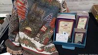 Woven village scene blanket with houses, trees, and snow covering, alongside the boxed set of Department 56 Heritage Village books in purple boxes.