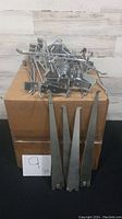 Box containing over 30 silver tone metal display brackets including several long flat brackets with hooked ends lying in front of the box