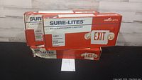 Three boxes of Sure-Lites exit and emergency lights stacked, showing general packaging and quantity.