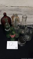 Photo showing all eight glass bottles arranged on black surface including vintage Coca-Cola bottles and various clear and brown glass bottles.