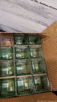 Top-down view of 12 green glass votive holders packed in a cardboard box, showing uniform size and color.