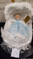 Porcelain doll dressed in blue lace dress, lying in white lace and satin cradle with certificate card attached.