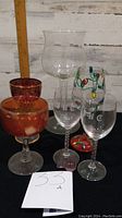Six miscellaneous glasses displayed on black surface with white wooden background, including different shapes and colors.