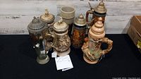 Wide view of all 7 decorative beer steins arranged on table to show variety and size