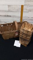 Photo showing a vintage wooden blueberry basket with handle alongside a stack of 16 square wooden baskets.