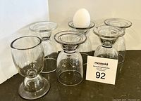 Six clear glass antique egg cups arranged with one holding an egg, showing flared rims and wide bases; some rims have chips due to age and wear.