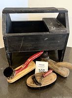 Photo showing wooden shoe shine box and assorted brushes arranged below it.
