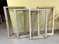 Photo showing four antique wooden window frames leaning against a yellow wall. Each frame includes glass panes; one has a circular etched design on the glass. Frames exhibit worn paint and aged wood surfaces, emphasizing their solid, heavy construction.