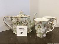 Porcelain tea pot and two mugs with white rose floral pattern and gold accents shown from the front