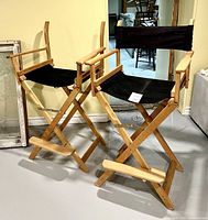 Photo showing two wooden directors chairs with black fabric seats and backs. One chair has torn or missing fabric on the backrest.