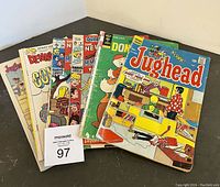 Photo showing 8 vintage comic books stacked fanned out, featuring Jughead, Dennis the Menace, and others with covers showing colorful comic scenes and titles.