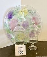 Two clear vintage apothecary jars with lids on a footed base in front of a large round glass plate decorated with pastel tulips in purple, yellow, and pink.