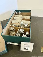 Box containing multiple pairs of salt and pepper shakers, separated with cardboard dividers, showing variety of styles and metal screw tops.