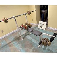 Weight bench setup shown with barbell loaded with plates on rack, dumbbells on bench, and attached leg developer with foam rollers.