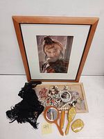Photo of the framed signed and numbered portrait 'Turksman Girl' by Gregory J Lawler resting above various vintage costume jewelry pieces including shells, stones, rosary beads, black lace scarf, and vintage vanity items like handheld mirrors and hairbrush.