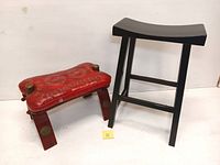 Two stools side by side, one red cushioned camel saddle seat with wooden legs and brass accents, and one black lacquered wood bar stool with curved seat and crossbar footrest.