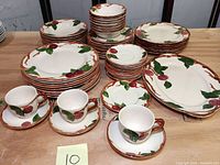 Full overview of dinnerware including stacked plates, bowls, platters, and front view of teacups with saucers showing the apple pattern and brown rim details