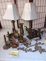 Photo showing two brass lamps with white shades, multiple candle holders, an elephant figurine, a vase, and assorted brass decor pieces arranged on a table.