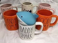 Six oversized mugs arranged in two rows: four Rae Dunn message mugs 'Mother Of the Bride', 'Bridesmaid', 'Food Coma', 'Candy Coma', one 'Daddy Shark' mug with blue shark lid in front and one Newport RI stein.