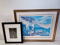 Full view of both framed artworks, including the colorful beach house print in a decorative gold frame and the railroad bridge photo in a black frame.