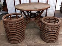 Gallery image showing full view of round table with woven rattan top and two cylindrical bamboo rattan stands, all placed on wood floor with folding screen background.