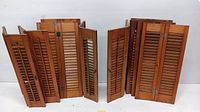Wide view of 12 wooden indoor shutters with slatted louvers and center hinges, grouped and standing upright.