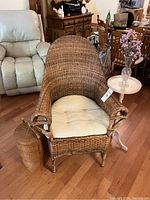 Wicker chair with cushion, small round side table with vase and flowers on top, wicker basket beside chair on wood floor.