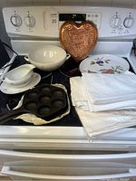 Photo showing the kitchenware and cast iron items arranged on a stove, including grav boat, bowl, copper wall hanging, plate, and linens.