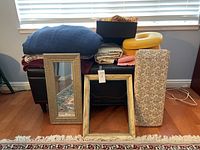 Photo showing chest with blankets, fabrics, yellow neck cushion, two framed mirrors, and floral fabric item in front.