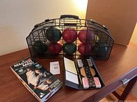 Game bundle items on a table in natural indoor lighting, showing a bocce ball set at back center, Mastermind game box on left, and black card case with decks of cards on right.