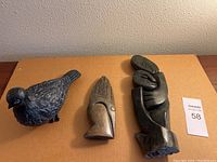 Three heavy sculptures on cardboard: a black detailed bird, a stylized bird head, and an abstract embracing figure sculpture.