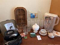 Photo showing an assortment of household items including glassware, ceramic tea set, wooden display case, plastic card holder, and a glass pitcher arranged on a table near a wall corner.