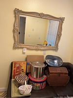 Photo showing large ornate mirror mounted on wall above assortment of home items on black bench including cooking pot, boxes and baskets