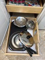 Kitchen drawer open showing stainless steel pots, pans, metal mixing bowl, grill pan, and glass lid.