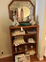 Wide view of wooden shelf holding folded linens and various decorative items, with octagonal floral etched wooden mirror hanging above it. Two porcelain vases and green ceramic dish on top of shelf.