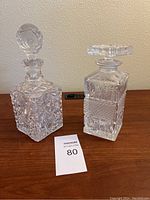 Two square vintage glass decanters with intricate cut glass designs displayed side by side including a round stopper and a flat wide stopper
