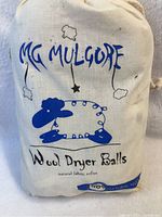 Cloth storage pouch showing MG Mulgore branding, sheep and star illustrations, labeled wool dryer balls, natural fabric softener.
