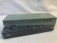 Two rectangular sharpening stones stacked with plastic wrapping on a light background. One stone is gray with 'Chef' branding and marked 400/1000 grit. The other stone is black with grip grooves.