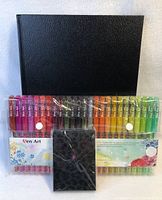 Two sets of colorful gel pens, a black sketchbook, and a black leopard print case arranged on a white background.