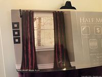 Packaging showing the curtains displayed hanging with three small framed pictures on the wall beside. Label indicates HALF MOON Prima Velvet Two Panels, plum color, solid velvet fabric.