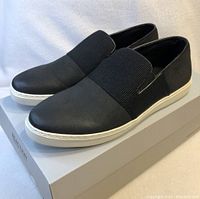 Side angled view of black Kenneth Cole slip-on loafers on box. Elastic band detail and white sole visible.