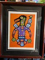Framed colorful print showing an abstract Thunderbird figure in orange background, signed by Christian Morrisseau