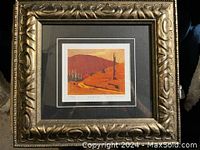 Photo of a framed decorative picture print with an ornate gold frame and black matte border, showing a colorful landscape artwork with orange and red tones.
