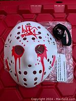 Front view of white Jason Voorhees hockey mask signed by Ari Lehman with red paint details and black adjustable strap on red surface